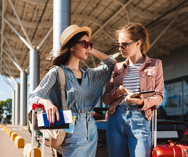Two female tourists with suitcases and passports standing outside a terminal reviewing their itinerary on a digital tablet, demonstrating seamless coordination and premium cruise guest experiences.