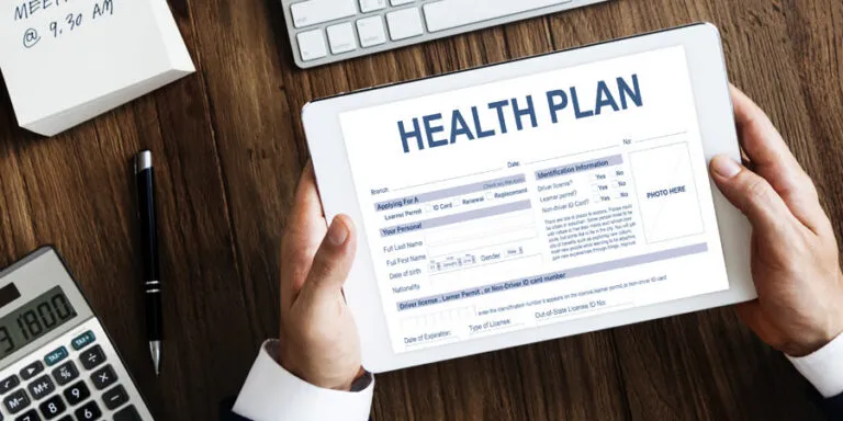 Hands holding a tablet showing a health plan form on a desk with a calculator